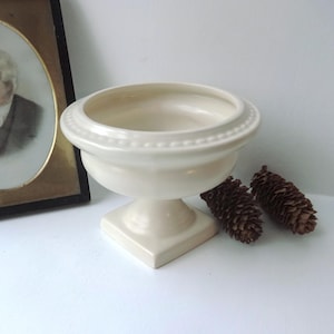 Może przedstawiać: Kremowa miska na cokole z ozdobnym, koralikowym brzegiem. Misa stoi na kwadratowej podstawie i obok dwóch brązowych szyszek. W tle znajduje się oprawiony portret mężczyzny.