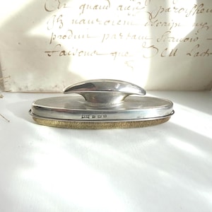 May include: A vintage silver powder compact with a domed top and a textured base. The compact has a silver rim and a gold-coloured, textured bottom. The background includes a handwritten document.