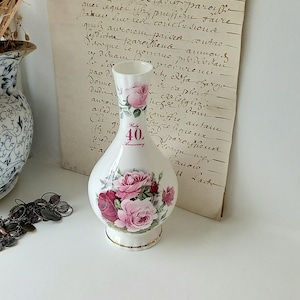 Può includere: Un vaso di ceramica bianco con un corpo bulboso e un collo stretto, ornato di rose rosa e rosse. Il vaso ha un bordo dorato e il testo "40th Anniversary". Un vaso floreale blu e bianco è sullo sfondo.