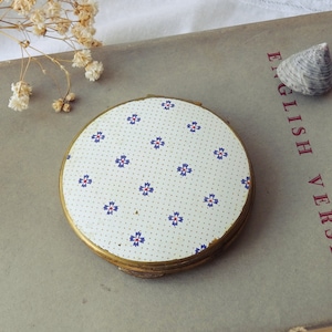 May include: A vintage compact mirror with a white dotted pattern and blue and red floral accents. The compact has a gold-toned metal frame and is resting on a book. Dried flowers and a seashell are also present.