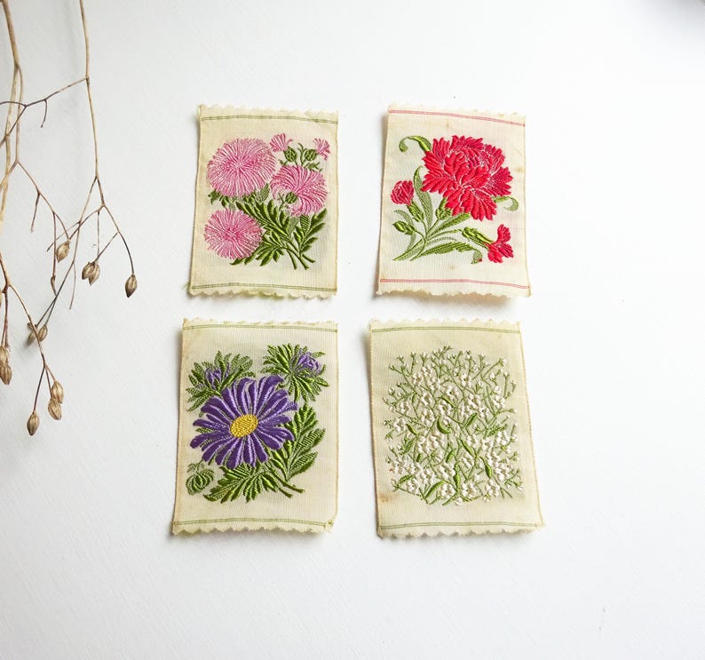 以下が含まれることがあります： 花柄の刺繍が施された布の正方形が4つ。正方形はクリーム色で、縁はギザギザになっている。デザインには、ピンクと白の花、赤い花、紫の花、白い花が含まれている。