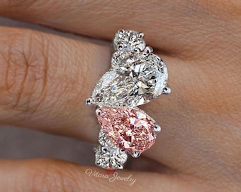 May include: A pear-shaped pink diamond ring with smaller white diamonds set in a silver band. The ring features three pear-shaped diamonds, one pink and two white, with smaller round diamonds surrounding them. The ring is set in a silver band and is a beautiful and elegant piece of jewelry.