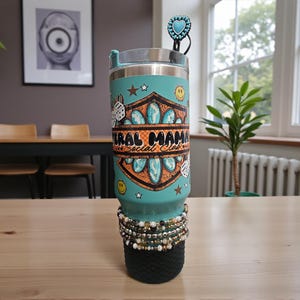 Puede incluir: Un vaso aislado turquesa con una banda decorativa con el texto "Liberal Mama Social Club". El vaso tiene una tapa plateada, un adorno de pajita turquesa y está adornado con pulseras. Una funda negra se encuentra en la parte inferior.