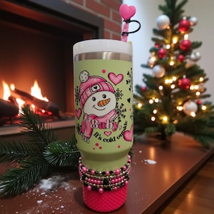 May include: A green and white insulated tumbler featuring a snowman design with a pink hat and scarf, hearts, and snowflakes. The tumbler has a pink base and is decorated with beads. A heart-shaped straw topper is visible.