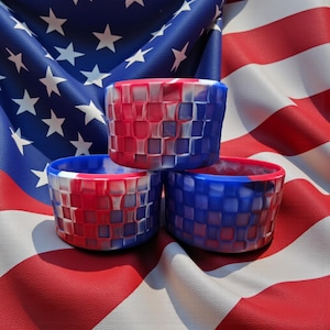 May include: Three woven-patterned silicone bowls in red, white, and blue, stacked on an American flag. The bowls have a cylindrical shape and a textured surface. The color scheme is patriotic, with the red, white, and blue representing the colors of the American flag.