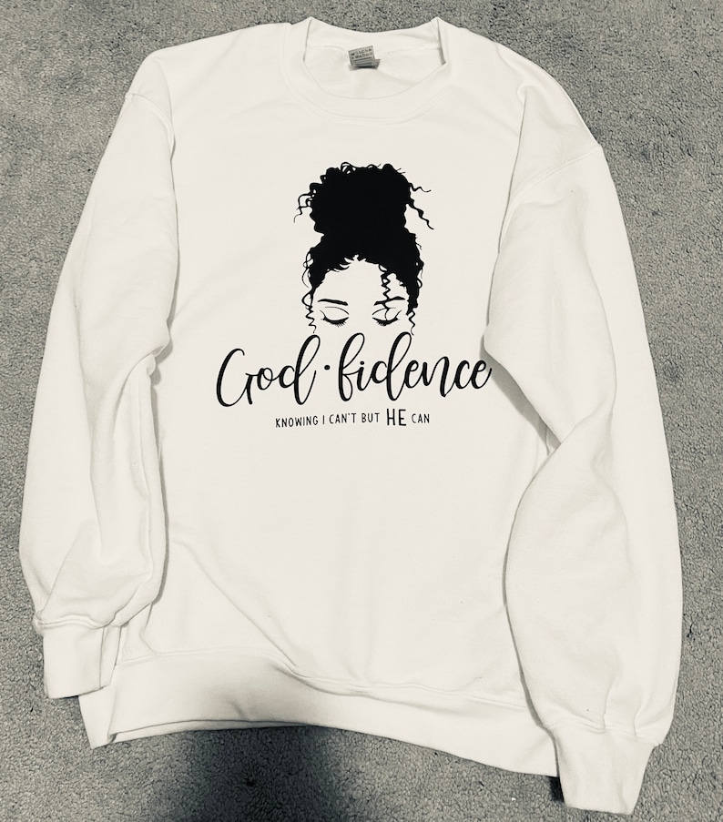 K&ouml;nnte beinhalten: Wei&szlig;er Pullover mit einem schwarzen Motiv einer Frau mit einem Dutt. Der Text "God-fidence" ist in Kursivschrift unter dem Bild geschrieben. Der Text "Knowing I can't but HE can" ist in Gro&szlig;buchstaben unter dem kursiv geschriebenen Text geschrieben.