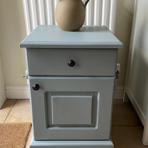 May include: A light blue painted wooden cabinet with a drawer and a door. A beige vase with green foliage sits on top. The cabinet has a dark knob on the drawer and door. The cabinet is 61 cm tall.