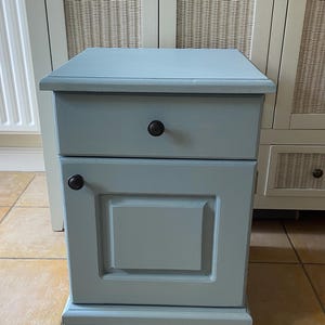 May include: A pale blue bedside cabinet with a drawer and a door, both with dark brown knobs. The cabinet is rectangular and has a painted matte finish. The dimensions are approximately 60cm high, 40cm wide, and 30cm deep.