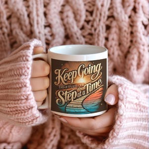 Może przedstawiać: Biały kubek ceramiczny z kolorowym wzorem graficznym. Wzór przedstawia zachód słońca ze ścieżką prowadzącą na szczyt wzgórza. Tekst "Keep Going Even If It's Just One Step At A Time" jest napisany czcionką w stylu vintage.