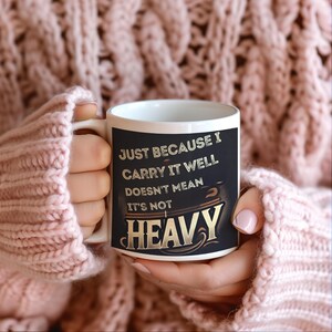 Może przedstawiać: Biały kubek ceramiczny z czarnym tłem i złotymi literami. Tekst brzmi "Just because I carry it well doesn't mean it's not heavy".