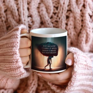 Może przedstawiać: Biały kubek ceramiczny z czarno-białym obrazkiem osoby niosącej duży kamień. Tekst na kubku brzmi "Just because I carry it well doesn't mean it's not heavy."
