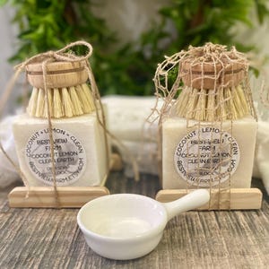 May include: Two square soap blocks with attached wooden scrub brushes, labeled "Coconut Lemon Clean" and "Best View Farm." A small white ceramic dish with a handle sits in front. The items are on a wooden surface.