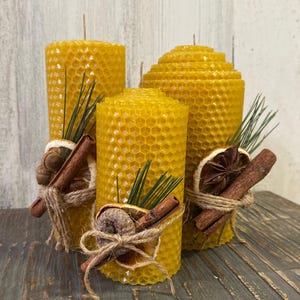 Puede incluir: Tres velas amarillas de cera de abeja con textura de panal, decoradas con palitos de canela, rodajas de naranja secas y cuerda. Las velas varían en altura y están dispuestas sobre una superficie de madera.
