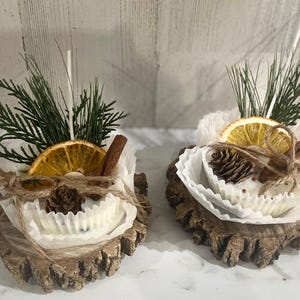 May include: Two white soy wax melts with a woodsy scent, decorated with dried orange slices, cinnamon sticks, pine sprigs, and pine cones. The melts are presented on rustic wood slices.