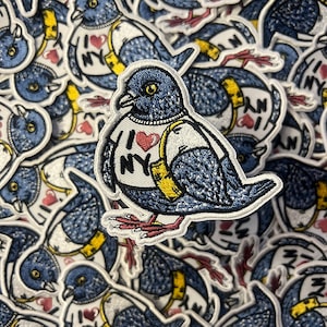 May include: A blue embroidered patch of a pigeon wearing a white shirt with the text "I ♥ NY" and a yellow bag. The pigeon has a red heart on its shirt and yellow feet.