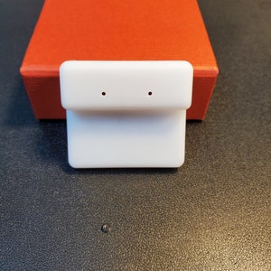 May include: A white earring display stand with two small holes, set in front of a textured red box. The stand is rectangular with a curved top. The background is a dark, textured surface.