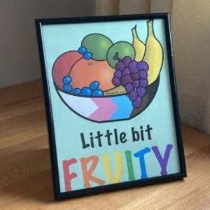Puede incluir: Una impresión enmarcada con un fondo verde que presenta un tazón de fruta con un melocotón, uvas, plátanos y manzanas verdes. El tazón está decorado con una bandera del orgullo transgénero. El texto "little bit FRUITY" está escrito debajo del tazón en colores arcoíris.
