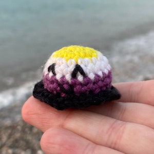 Puede incluir: Un pequeño gorro de crochet en los colores de la bandera del orgullo no binario. El gorro es amarillo, blanco, morado y negro.