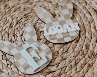 May include: Two wooden bunny-shaped ornaments with a checkered pattern. One ornament has the name "Logan" written on it in white letters. The other ornament has the letter "E" written on it in light blue letters.