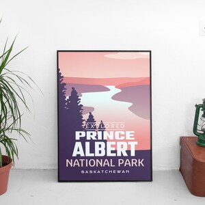 May include: A framed poster with a landscape design featuring a river and trees, with text that reads "EXPLORED PRINCE ALBERT NATIONAL PARK SASKATCHEWAN". A green lantern and a brown suitcase are next to the poster. A potted plant is on the left.