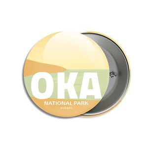 May include: A round button with a glossy finish, featuring the word "OKA" in white block letters. Below, it reads "NATIONAL PARK QUEBEC." The design includes a landscape in shades of yellow, green, and brown.