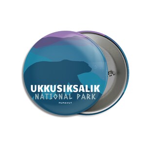 May include: A circular badge with a bear silhouette set against a blue and purple backdrop. The text "UKKUSIKSAALIK NATIONAL PARK" is in white, with "NUNAVUT" underneath. The badge has a metal pin fastening on the reverse.