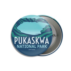 Puede incluir: Un botón redondo con una ilustración paisajística del Parque Nacional Pukaskwa en Ontario, Canadá. El diseño presenta un lago y montañas en tonos azules y verdes, con el nombre del parque en texto blanco. El botón mide unos 5,7 cm de diámetro.