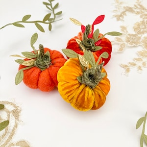 May include: Three felt pumpkins in orange, red and yellow with green stems and leaves. The pumpkins are arranged on a white background.