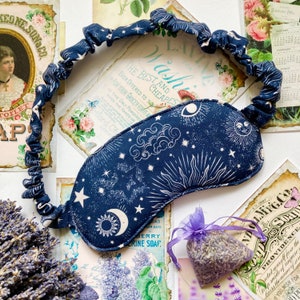 May include: A blue sleep mask with a white celestial print featuring stars, moons, and suns. The mask has an elastic band for a comfortable fit.