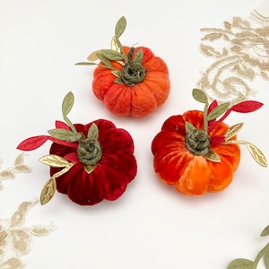 May include: Three velvet pumpkins in different colours: red, orange, and a lighter orange. The pumpkins are decorated with green leaves and gold accents.