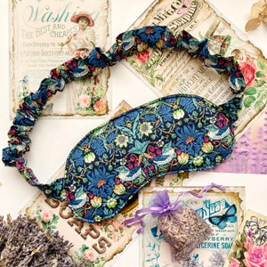 May include: A blue and green floral eye mask with a ruffled edge. The mask is made of fabric with a floral pattern and has a white background. The mask is lying on a white surface with vintage-style paper and lavender flowers.