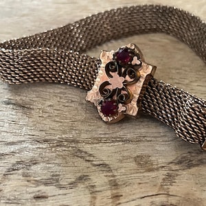 May include: A gold mesh bracelet with a decorative clasp featuring three red gemstones. The clasp is ornate and has a floral design.
