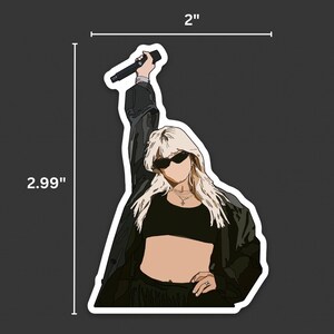 May include: A sticker of a person with blonde hair wearing sunglasses and a black crop top, holding a microphone. The person is wearing a black jacket and black pants. The sticker is 2 inches by 2.99 inches.
