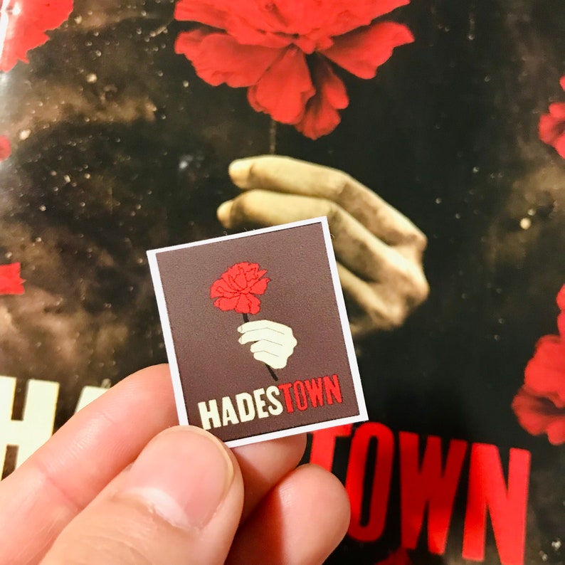 May include: A sticker with a brown background and a red carnation being held by a white hand. The text "Hadestown" is printed in white letters on the sticker.