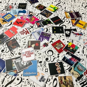 May include: A collection of colorful square stickers featuring the names of various Broadway musicals, including "Wicked", "Chicago", "Annie", "The Music Man", "Hadestown", "The Great Comet", "Suffs", "Kimberly Akimbo", "Mrs. Doubtfire", "Go-Go Dolls", and "Mamma Mia".