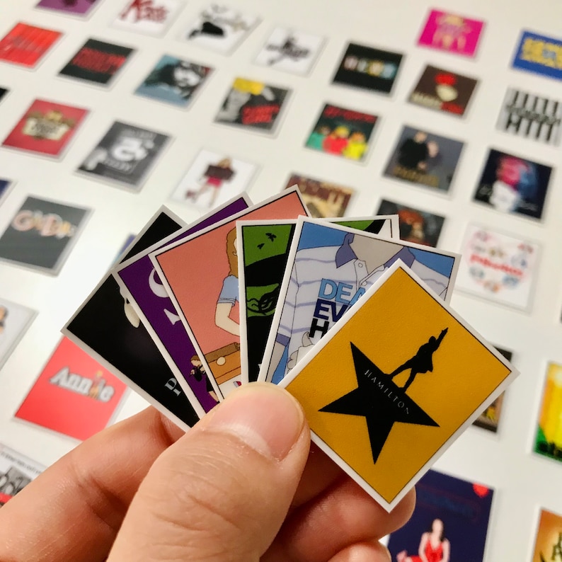 May include: A collection of colorful square stickers featuring various Broadway show logos, including "Annie", "Dear Evan Hansen", "Hamilton", and "The Greatest Showman".