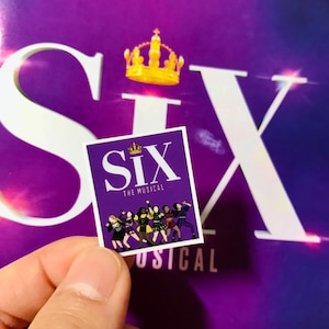 May include: A purple sticker with the words "Six The Musical" and a crown above the word "Six". There are six women in colorful outfits standing in a row on the sticker.