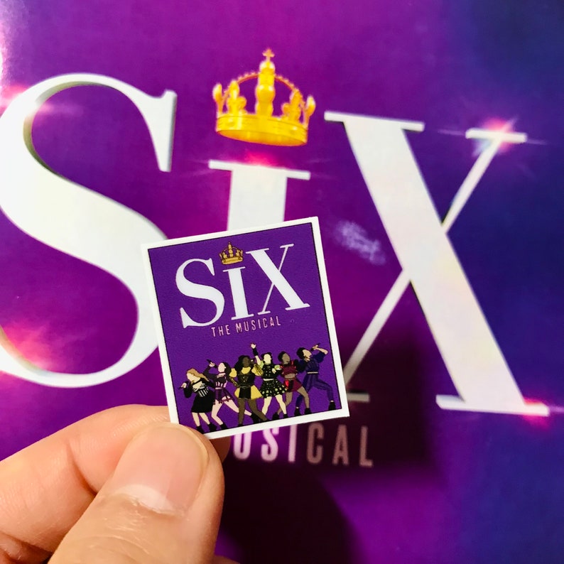 May include: A purple sticker with the words "Six The Musical" and a crown above the word "Six". There are six women in colorful outfits standing in a row on the sticker.