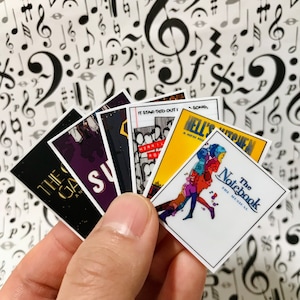May include: A collection of six colorful musical show posters. The posters are held in a hand. The posters feature titles like "The Game", "Super", "It Started Out I", "Hell's Kitchen", and "The Notebook".