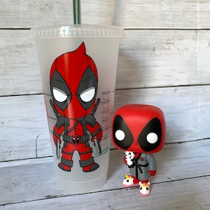 以下が含まれることがあります： 透明なプラスチック製の再利用可能なカップで、赤と黒のカートゥーンのデッドプールキャラクターのデザインが施されています。カップの隣には、より小さな赤と黒のデッドプールフィギュアが立っています。