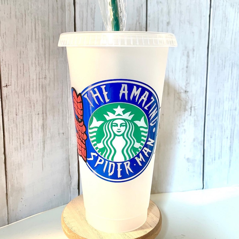 May include: A white reusable cup with a blue and green Starbucks logo that says "The Amazing Spider-Man". The cup has a red and blue Spider-Man design on the side.