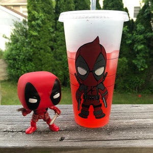 以下が含まれることがあります： 赤い液体が入った透明なプラスチックカップに、赤と黒のデッドプールキャラクターがプリントされています。手前に、赤と黒の小さなデッドプールフィギュアがあります。