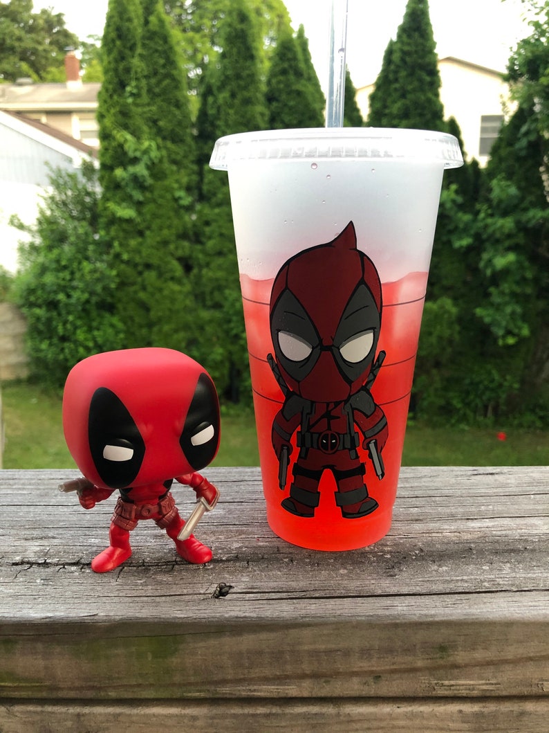以下が含まれることがあります： 赤い液体が入った透明なプラスチックカップに、赤と黒のデッドプールキャラクターがプリントされています。手前に、赤と黒の小さなデッドプールフィギュアがあります。
