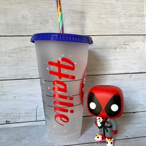 以下が含まれることがあります： 青い蓋と赤い「コーヒー」のデザインが施された透明なプラスチック製の再利用可能なカップ。カップは白いグリッターで飾られています。カップの隣には、赤と黒のデッドプールフィギュアが立っています。