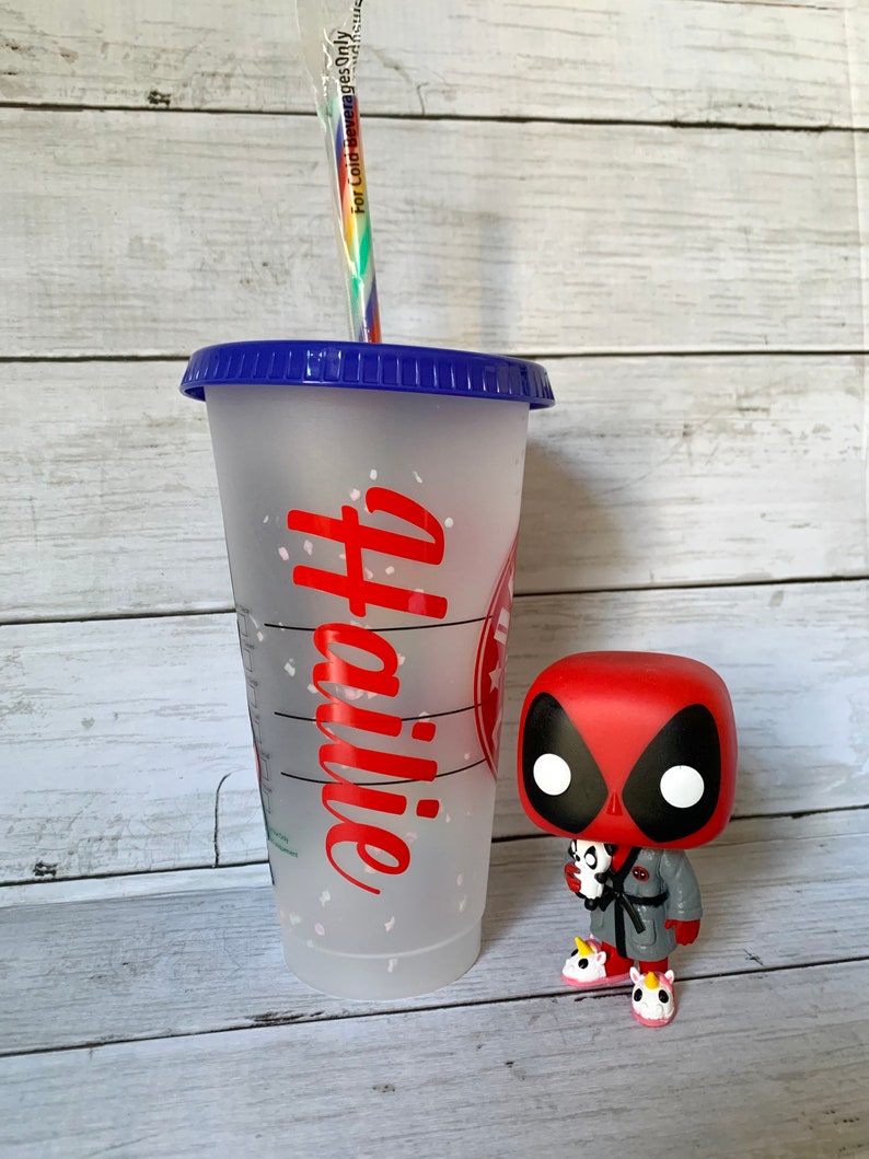 以下が含まれることがあります： 青い蓋と赤い「コーヒー」のデザインが施された透明なプラスチック製の再利用可能なカップ。カップは白いグリッターで飾られています。カップの隣には、赤と黒のデッドプールフィギュアが立っています。