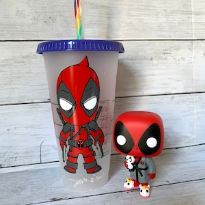 以下が含まれることがあります： 青い蓋と赤と黒のカートゥーンのデッドプールデザインが施された透明なプラスチック製のタンブラー。タンブラーの隣には、赤と黒のデッドプールの小さなフィギュアが立っています。フィギュアは灰色のローブを着て、ピンクのユニコーンのスリッパを履いています。