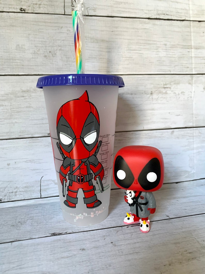 以下が含まれることがあります： 青い蓋と赤と黒のカートゥーンのデッドプールデザインが施された透明なプラスチック製のタンブラー。タンブラーの隣には、赤と黒のデッドプールの小さなフィギュアが立っています。フィギュアは灰色のローブを着て、ピンクのユニコーンのスリッパを履いています。