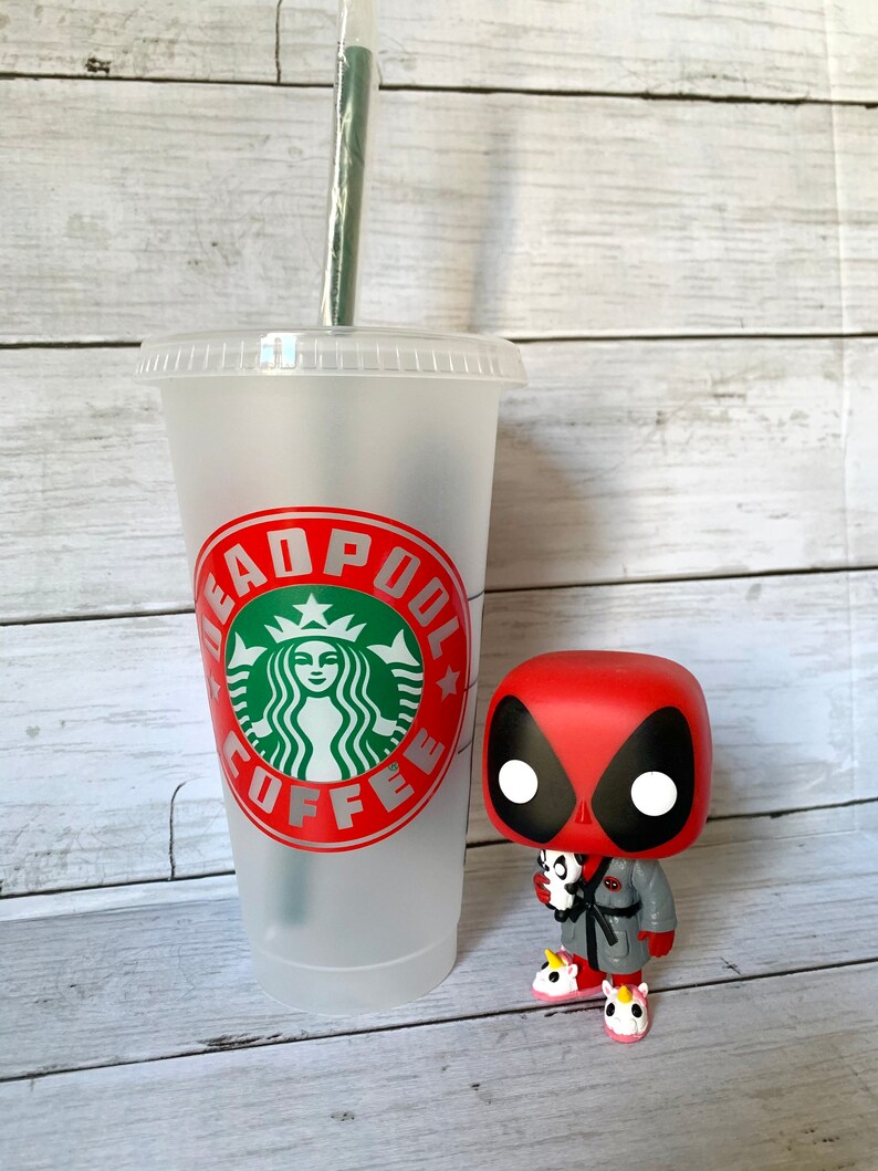 以下が含まれることがあります： 赤い緑のデッドプールコーヒーのロゴが入った白い再利用可能なコーヒーカップ。グレーのローブとユニコーンのスリッパを履いたデッドプールのファンコポップフィギュアがカップの隣に立っています。