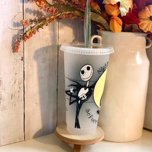 May include: A clear plastic tumbler with a design of Jack Skellington from The Nightmare Before Christmas. The tumbler has a lid and a green straw. The words "Nightmare Before" are printed on the cup. The cup is on a small wooden stand.