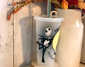 May include: A clear plastic tumbler featuring a design of Jack Skellington from The Nightmare Before Christmas. The tumbler includes a lid and a green straw. The words "Nightmare Before" are printed on the cup. The cup is displayed on a small wooden stand.